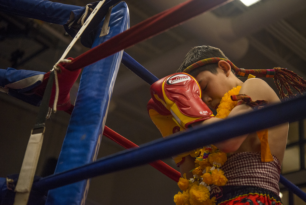 A boy in traditional Muay Thai ceremony, wearing a mongkhon headband and marigold garland, eyes closed in prayer before a fight